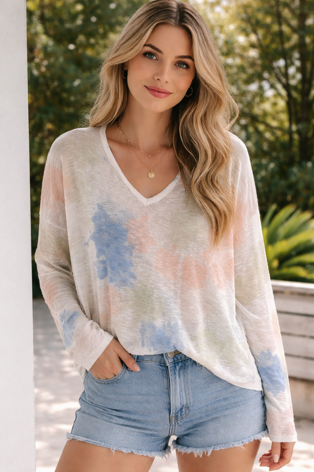 Woman wearing pastel tie dye V-neck lightweight sweater with soft blue, peach and sage tones paired with denim shorts.