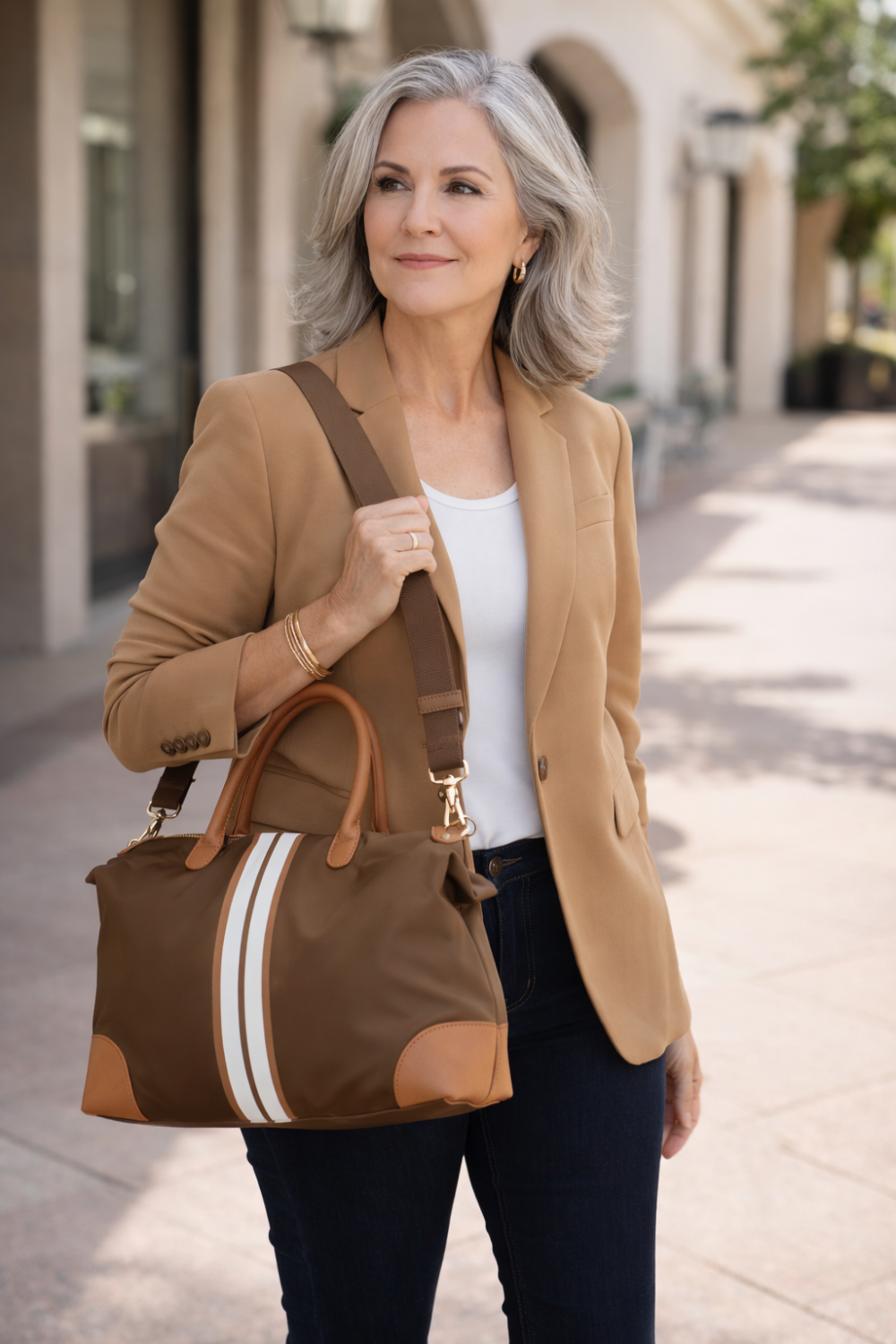 Confident woman age 45-60 wearing a camel blazer and jeans carrying a luxury brown stripe weekender tote bag in an upscale outdoor setting.