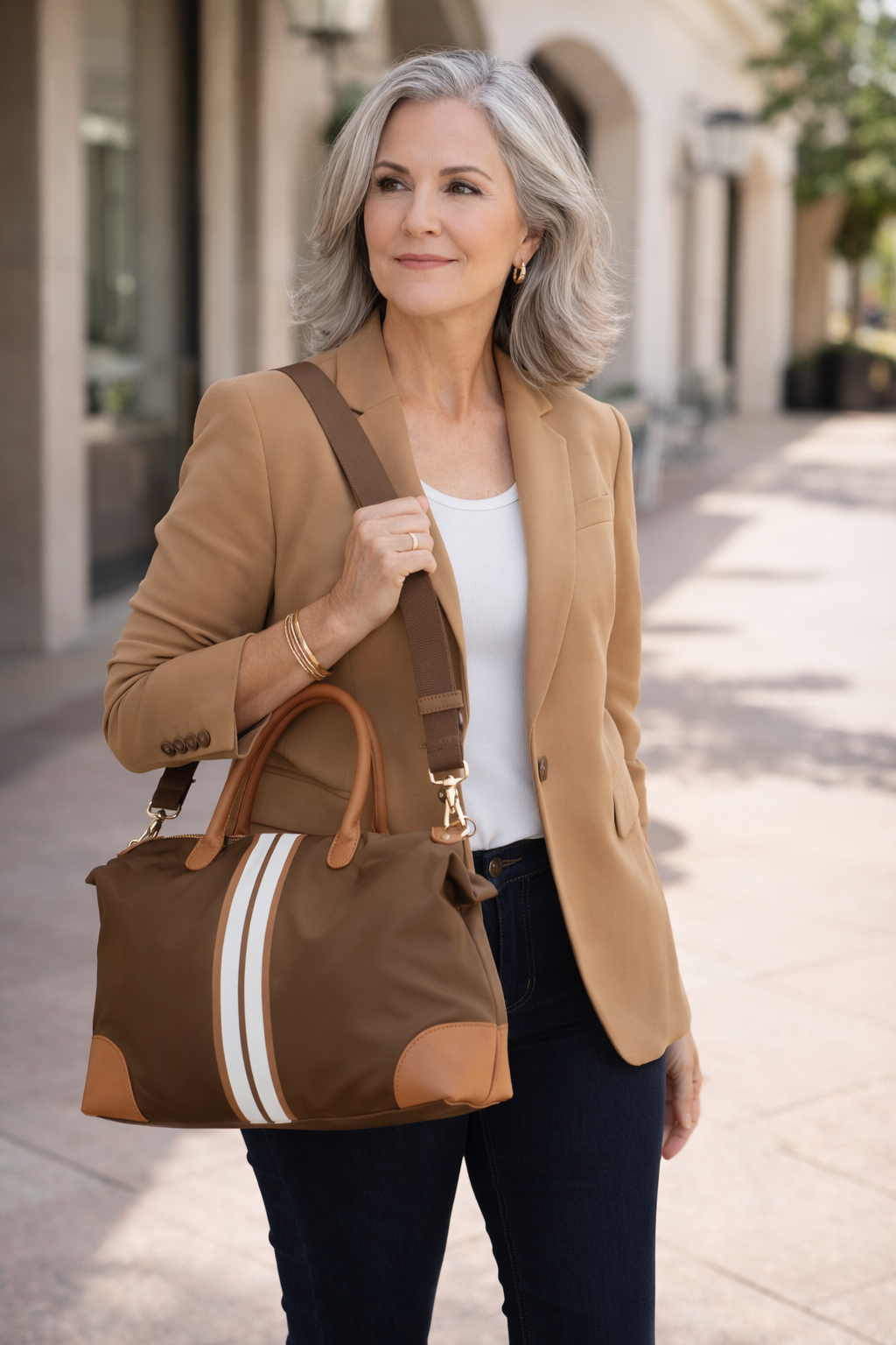 Confident woman age 45-60 wearing a camel blazer and jeans carrying a luxury brown stripe weekender tote bag in an upscale outdoor setting.